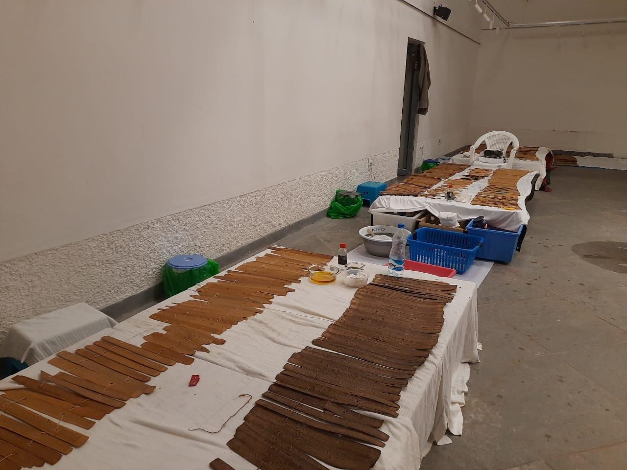 A row of long tables covered in white cloth, with palm leaves laid out in arrays and bowls of oil alongside, showing batch processing of many manuscripts at once.